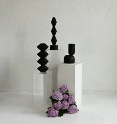 Three black sculptural objects on white pedestals with a cluster of purple hydrangeas at the base.