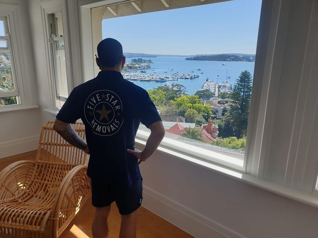 Man in work shirt gazes out the window at a scenic harbor. Calm blue water and boats fill the view outside the window.