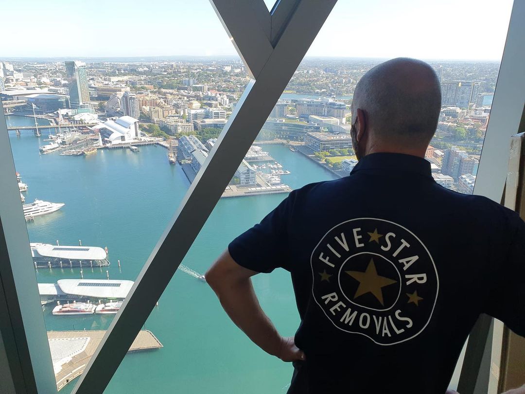 Man looking at the cityscape through a window; he's wearing a dark blue polo shirt with the words 'FIVE STAR REMOVALS' on the back.