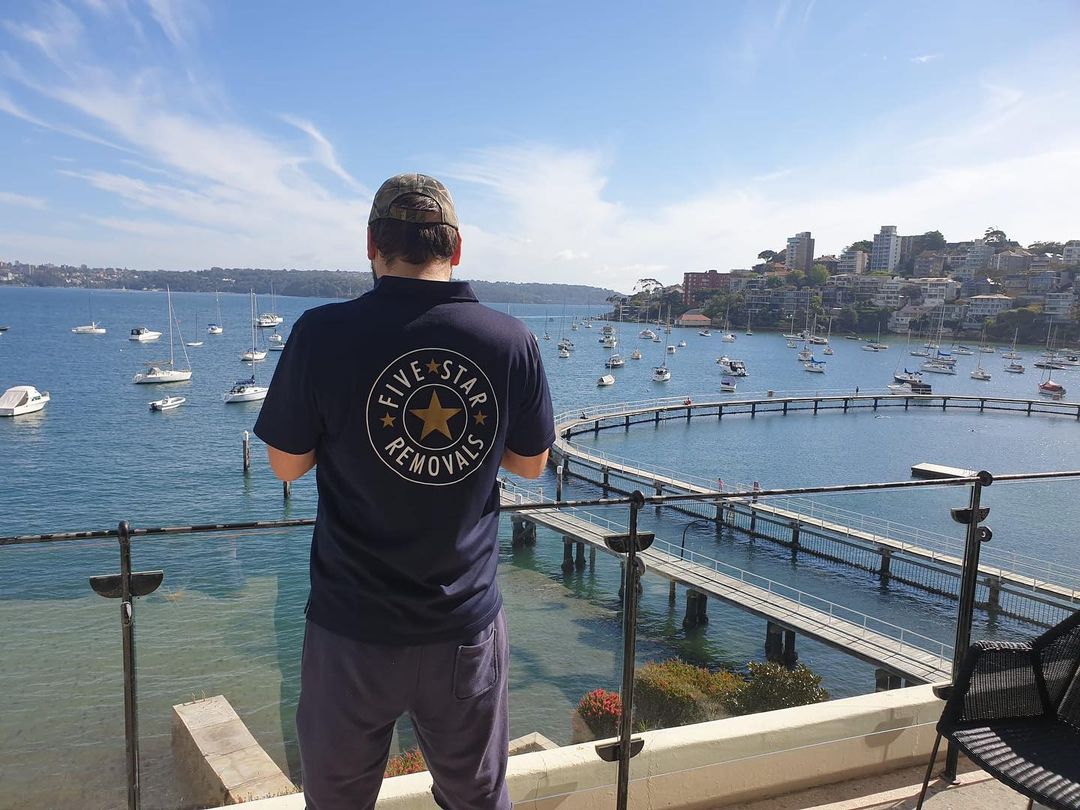 Man overlooking Sydney harbour from a balcony, wearing a shirt with a Five Star Removals logo, bridge, boats, and buildings.