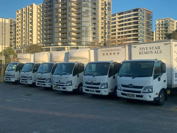 Line of white Five Star Removal trucks parked in a lot, with tall apartment buildings visible in the background under a clear sky.