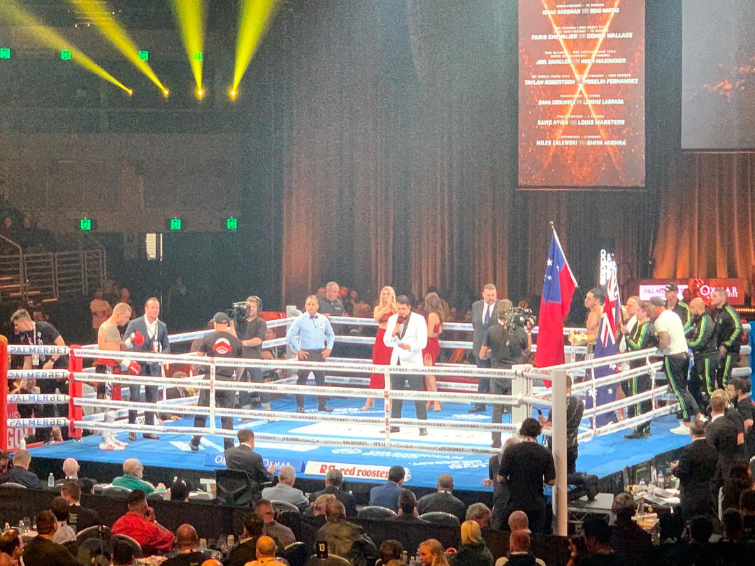 Boxing match underway in a brightly lit arena, featuring boxers in the ring with referees and corner teams, captured from an audience perspective.