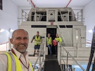 A selfie capturing a flight simulator bay with 4 people standing in front of the cockpit, wearing safety vests, in a bright room.