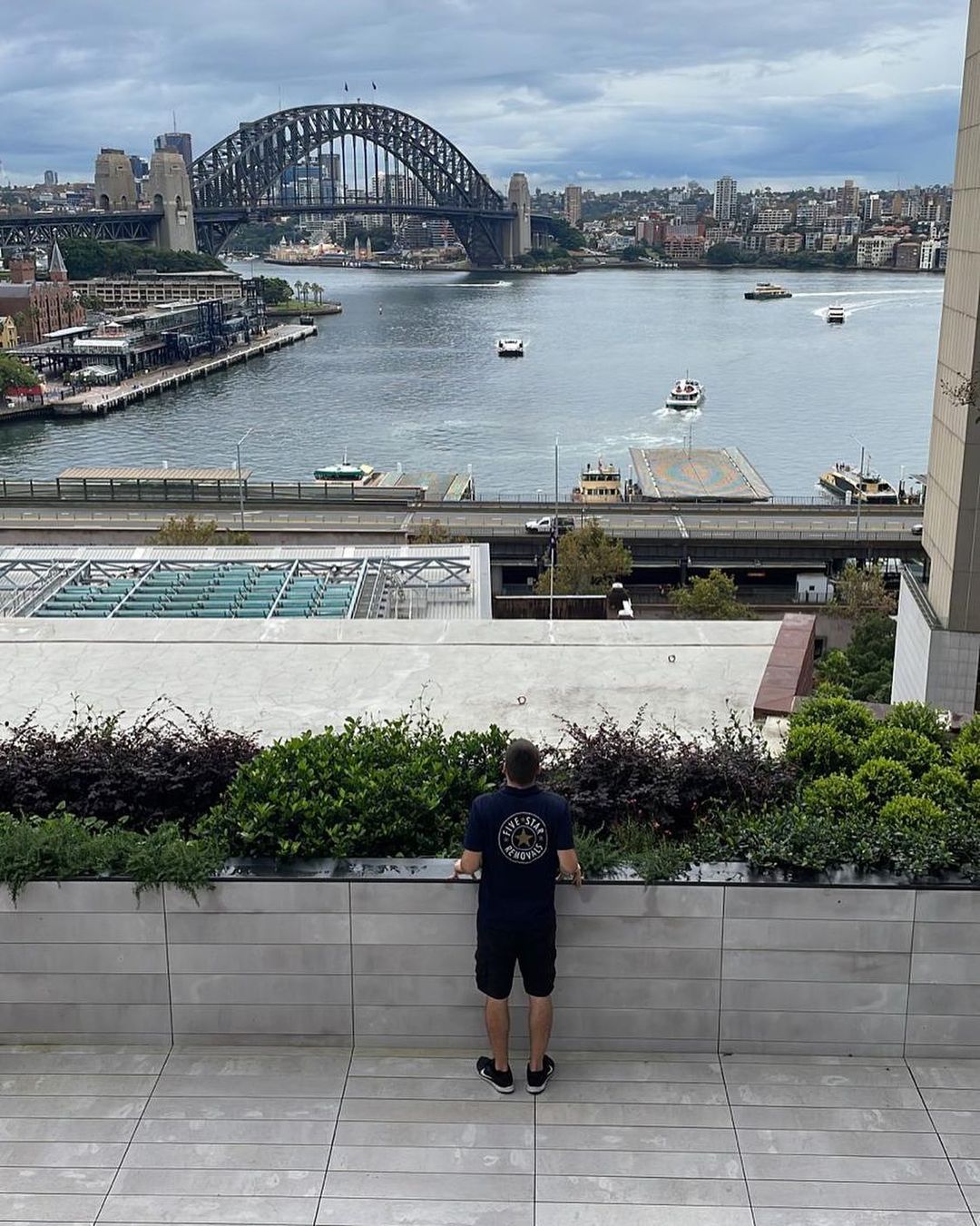 Man gazes at Sydney Harbour Bridge and city skyline from rooftop terrace. Watercraft float on the harbour in the daytime under cloudy skies.