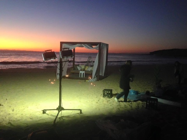 Beach setting with cabana-style bed, lighting, and people against a twilight sky. Sand, ocean, and stage lighting illuminate the scene.