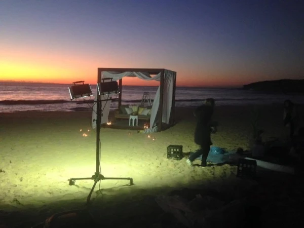 Beach setting with cabana-style bed, lighting, and people against a twilight sky. Sand, ocean, and stage lighting illuminate the scene.