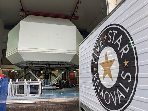 Indoor shot of a full flight simulator, featuring a white exterior, supported by machinery with 'Five Star Removals' logo truck partially visible.