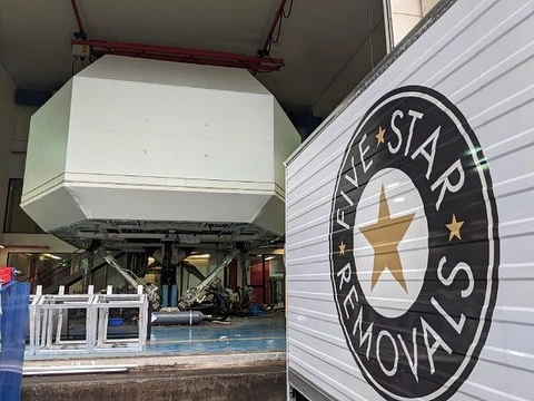Indoor shot of a full flight simulator, featuring a white exterior, supported by machinery with 'Five Star Removals' logo truck partially visible.
