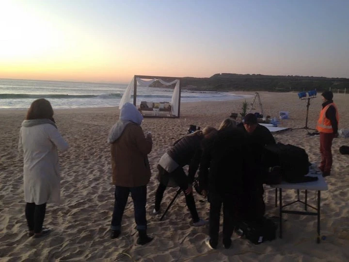 Beach photoshoot with a crew of people, lighting, and staging at sunset, capturing the scene with the ocean and horizon visible.