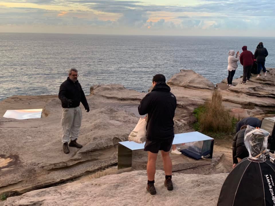 Outdoor photography shoot on a rocky coastline with five people, ocean view, photo equipment, and reflective panels.