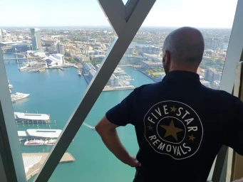 Man wearing a dark blue polo shirt with a gold star logo looking out a large window overlooking a coastal city harbour.