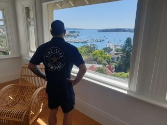 Man wearing a navy blue t-shirt with Five Star Removals logo standing by a large window looking out at a harbour view in Sydney