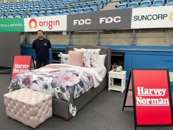Man standing next to a made bed set up on a carpeted floor in a stadium with corporate sponsor signs behind it.