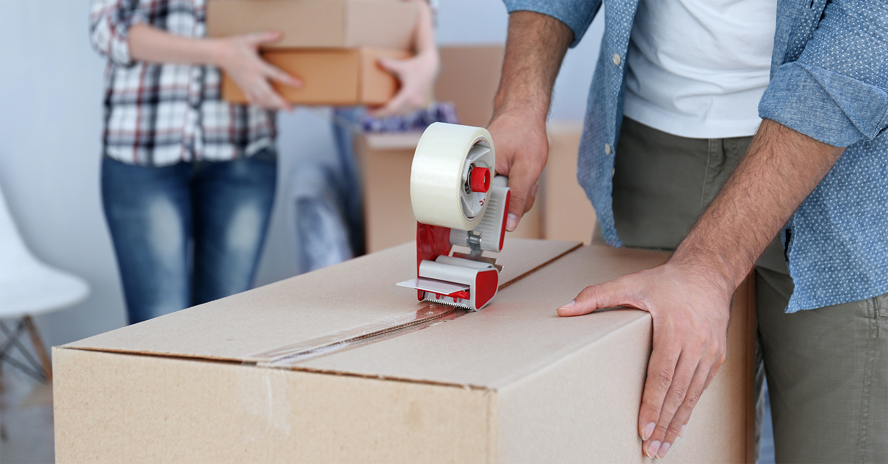 Adults packing boxes in a brightly lit indoor space, using a tape dispenser, preparing for relocation or shipping.