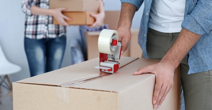 Adults packing boxes in a brightly lit indoor space, using a tape dispenser, preparing for relocation or shipping.