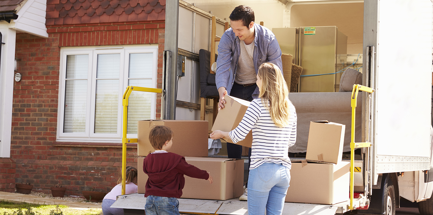 Family of four unloads cardboard boxes from moving truck outside a brick house on a sunny day, symbolizing relocation and new beginnings.