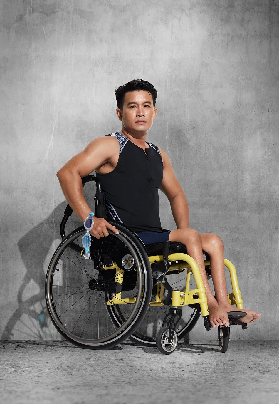 An adult male athlete with a disability is sitting in a yellow wheelchair in a studio environment, wearing a black top and holding goggles.