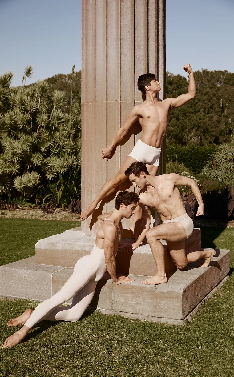 Three men in dance poses near a stone column in a grassy park setting, creating a sculptural tableau in outdoor lighting.