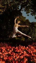 Male ballet dancer in a white leotard leaping gracefully over a bed of red flowers beneath lush green trees in Sydney.