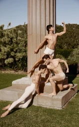 Three male dancers posing gracefully in white dancewear around a classical stone column outdoors on a sunny green lawn.