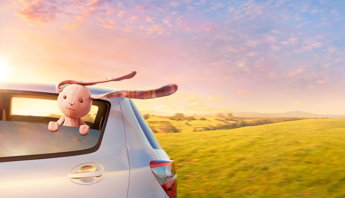 Stuffed bunny toy peers from a car window during a scenic drive, set against a backdrop of rolling green hills and a pastel sky.