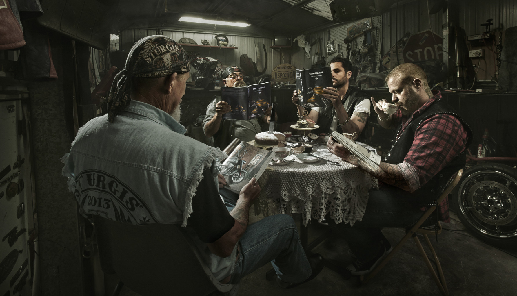 Five adults gather around a table set for tea and reading motorcycle magazines in a cluttered garage or workshop with dim lighting.