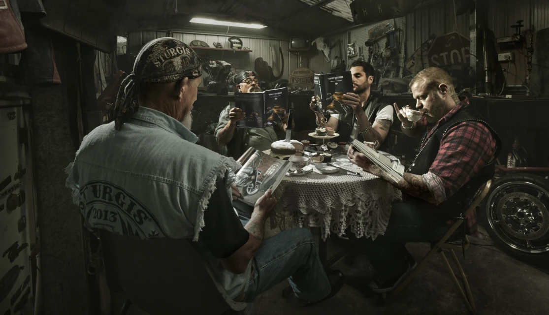Five adults gather around a table set for tea and reading motorcycle magazines in a cluttered garage or workshop with dim lighting.