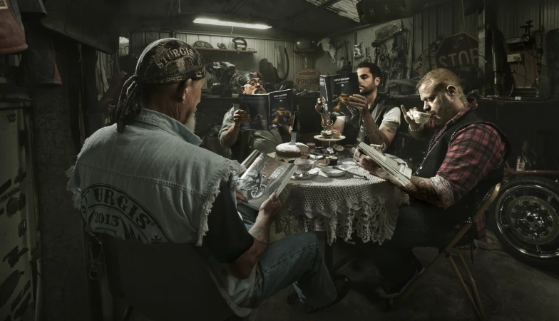 Five adults gather around a table set for tea and reading motorcycle magazines in a cluttered garage or workshop with dim lighting.