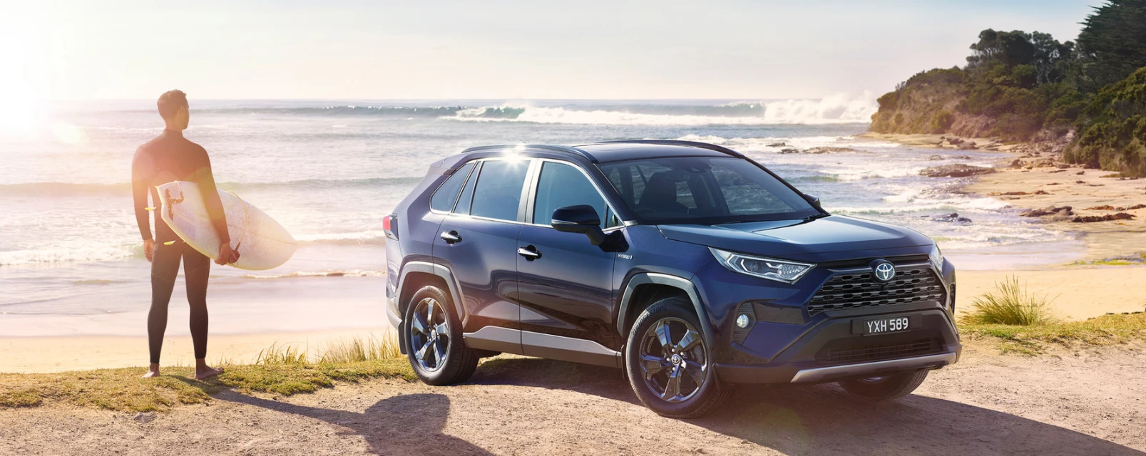Blue SUV parked on sandy beach with surfer walking towards ocean waves on a sunny day, showcasing automotive and travel lifestyle.