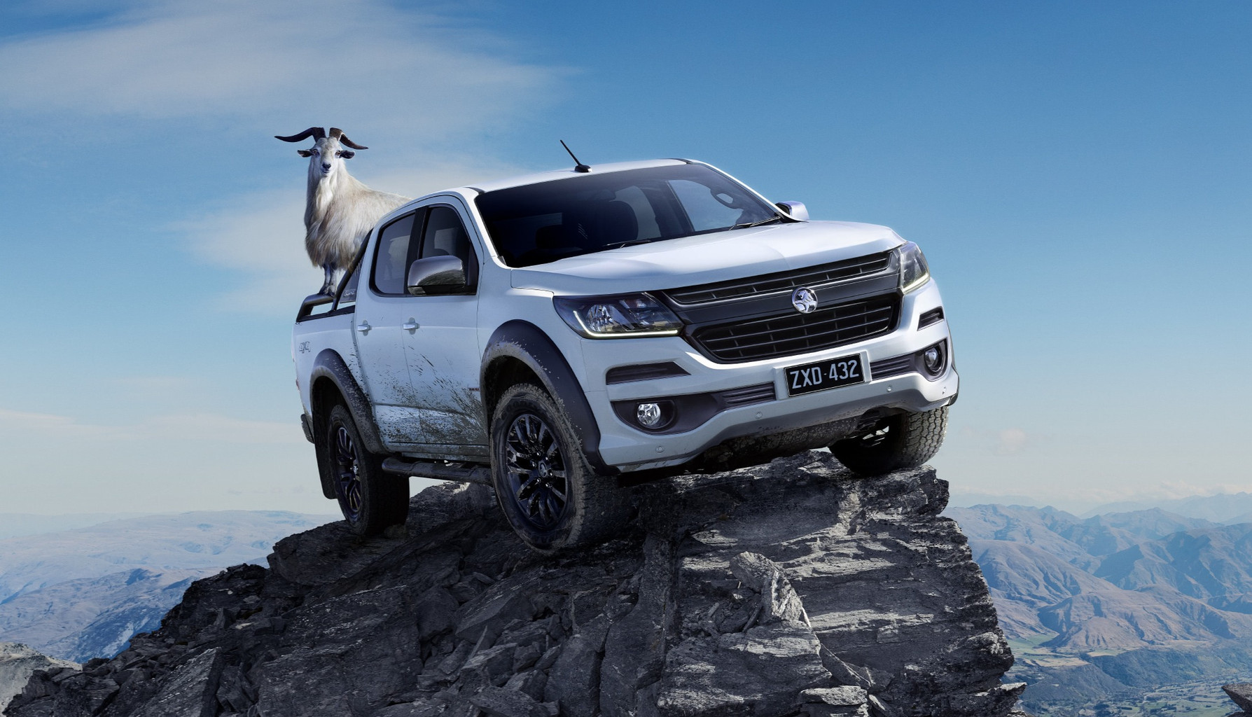 White pickup truck on a rocky mountain peak with a goat standing in the truck bed against a backdrop of mountains and blue sky.