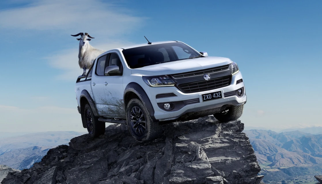 White pickup truck on a rocky mountain peak with a goat standing in the truck bed against a backdrop of mountains and blue sky.