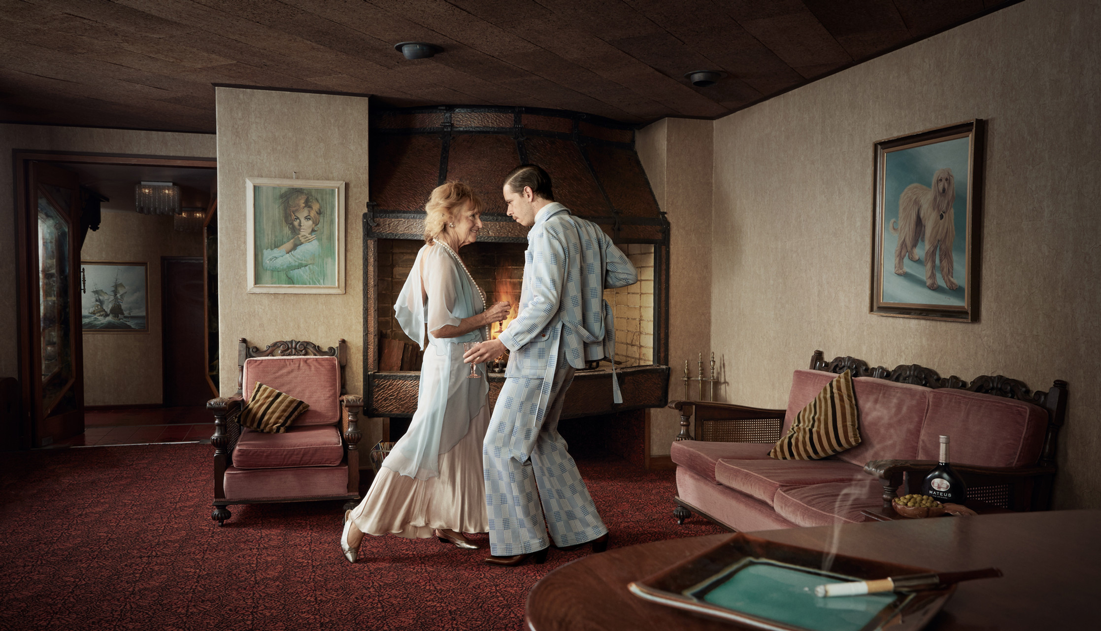 A mature couple in formal attire dances intimately inside a room with vintage decor, illuminated by soft lighting and warm color grading.