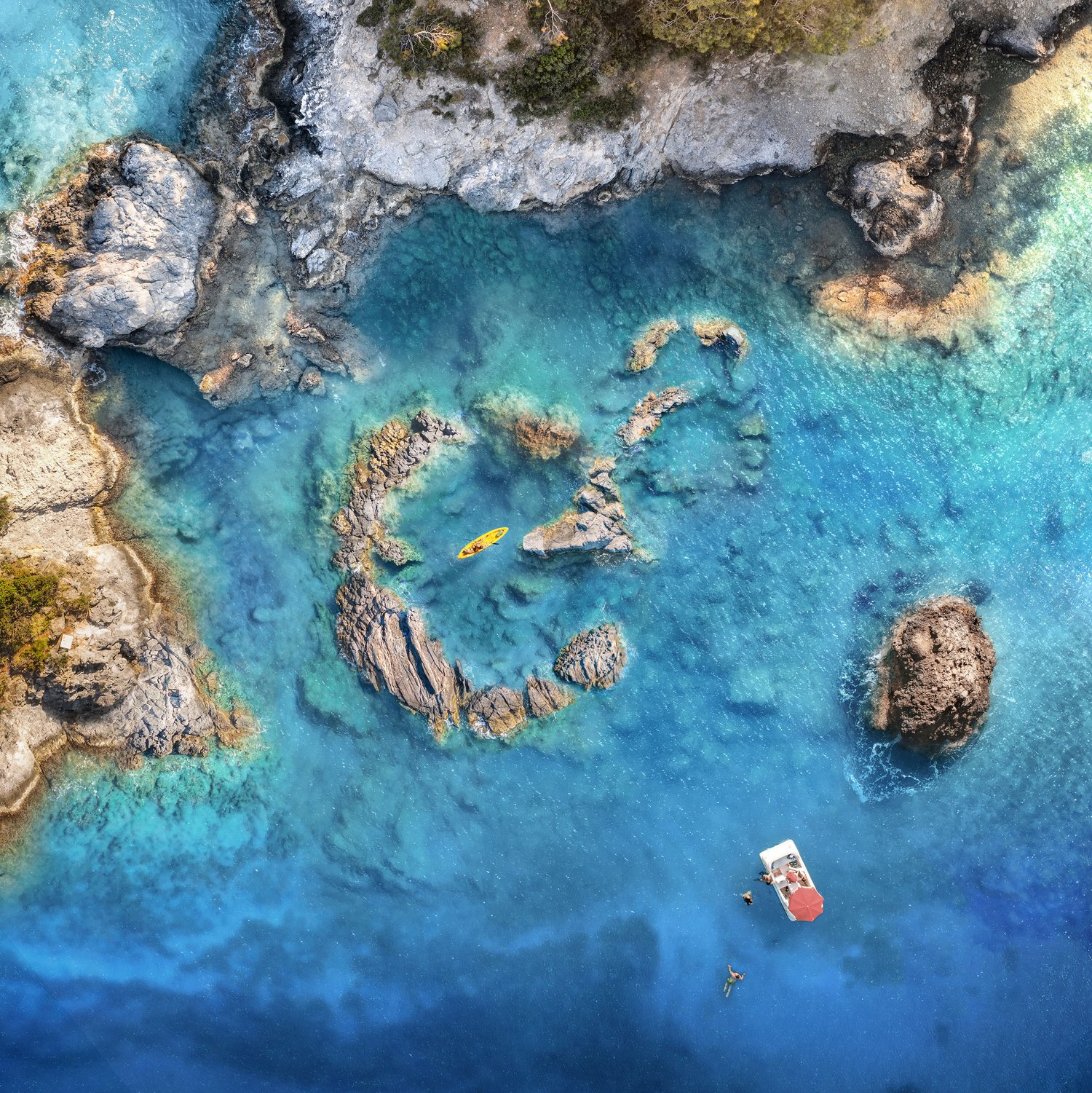 Aerial view of clear turquoise sea water with rocky coastline, a kayak, small boat, and people swimming, in a summer travel scene.