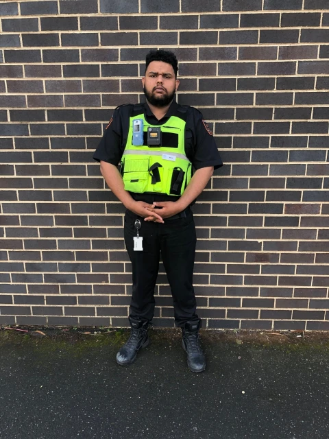 Standing portrait of adult male in a black uniform and neon yellow vest with a brick wall background on an overcast day.