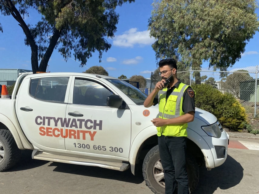 Security guard in a high-visibility vest talks on a walkie-talkie next to a security company truck in an outdoor environment during daytime.