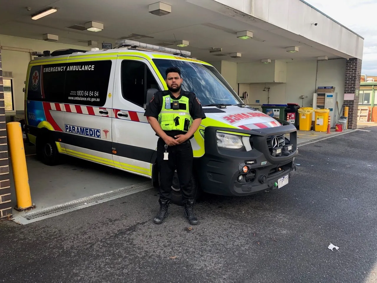 Paramedic stands in uniform in front of a white and yellow emergency ambulance outside a building, looking directly at the camera.