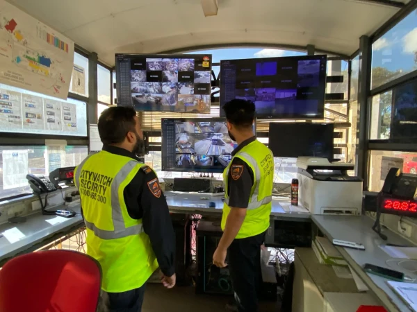 Two security guards in uniform stand inside a security watchtower with multiple screens displaying surveillance footage, monitoring the area.