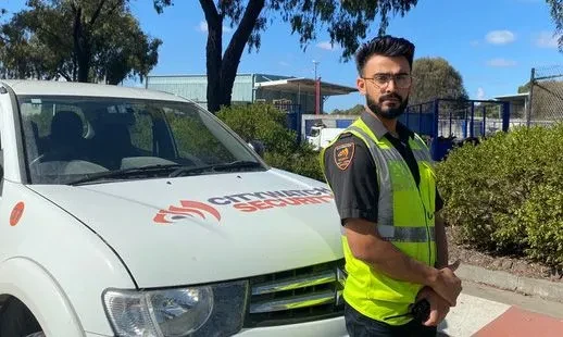 Adult male security guard in a hi-vis vest stands next to a white Citywatch Security truck outdoors on a sunny day in an urban environment.