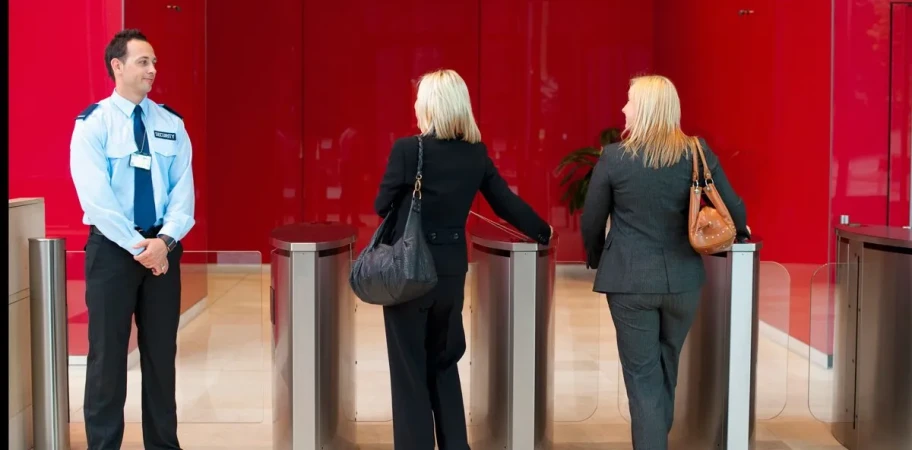 Two women enter a building through turnstiles as a security guard watches, set against a red wall in a modern office setting.