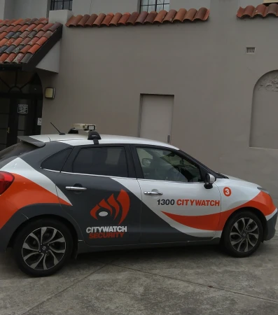 Citywatch Security car parked in front of a building with a red-tiled roof, featuring the company's logo and phone number.