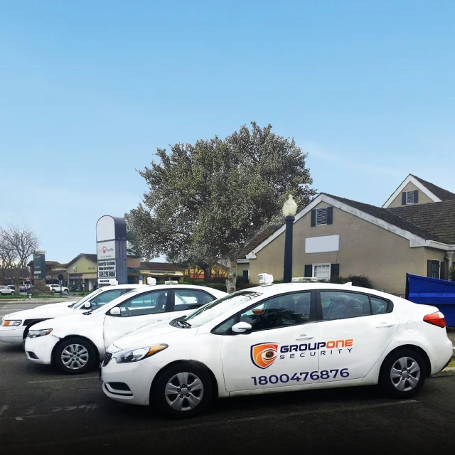 White cars parked outdoors with visible Group One Security logo, clear blue sky, and commercial buildings in the background during daytime.