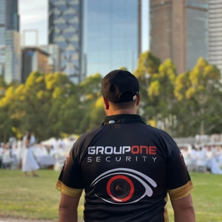 Security guard in black uniform with Group One Security logo on green lawn with buildings in background, viewed from behind.
