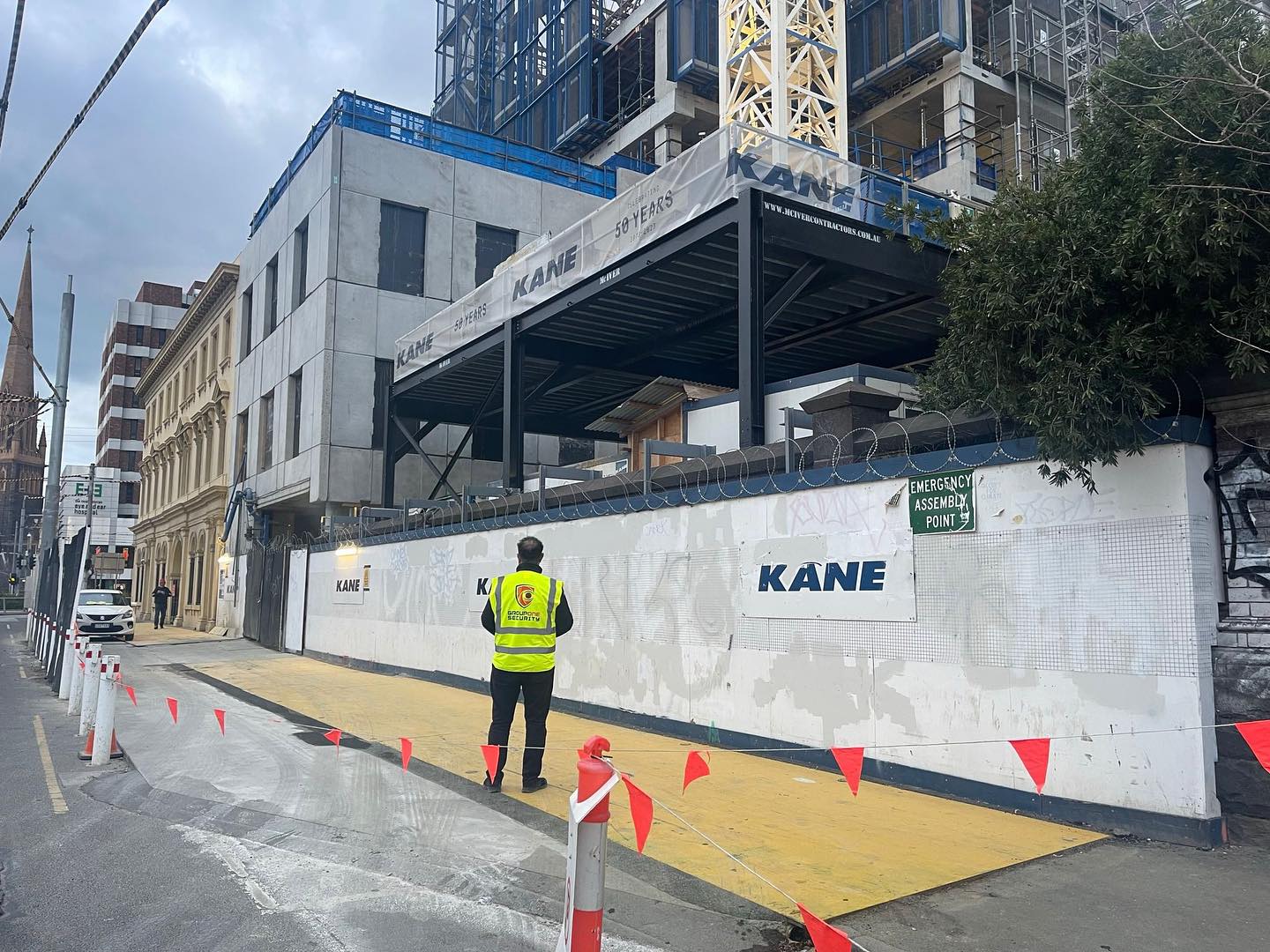 Construction worker in safety vest walks past new building under construction with a crane and old brick building along a city street.