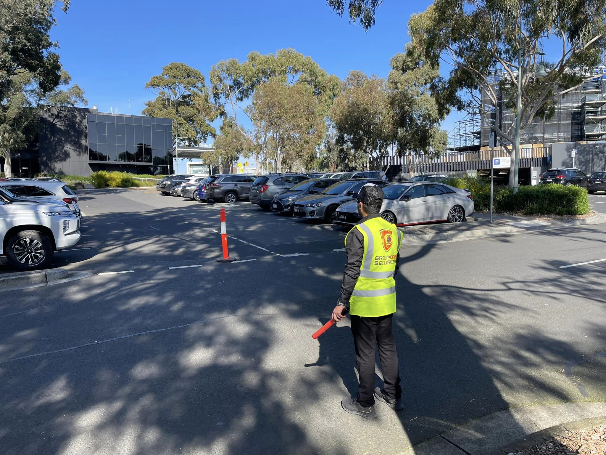 A parking attendant in a bright yellow vest stands in a parking lot full of cars on a sunny day, directing traffic with a red baton.