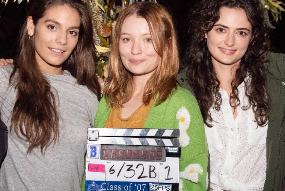 Three women pose indoors with a production slate showing production details. They are smiling and dressed casually in a well-lit scene.