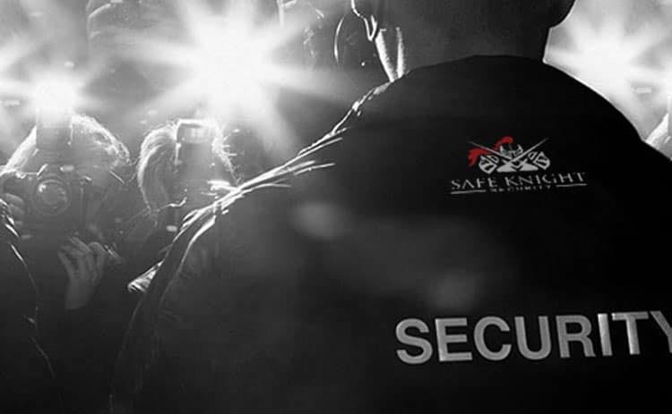 A security guard in a black jacket with 'SECURITY' printed on the back stands in front of a crowd of photographers with camera flashes.