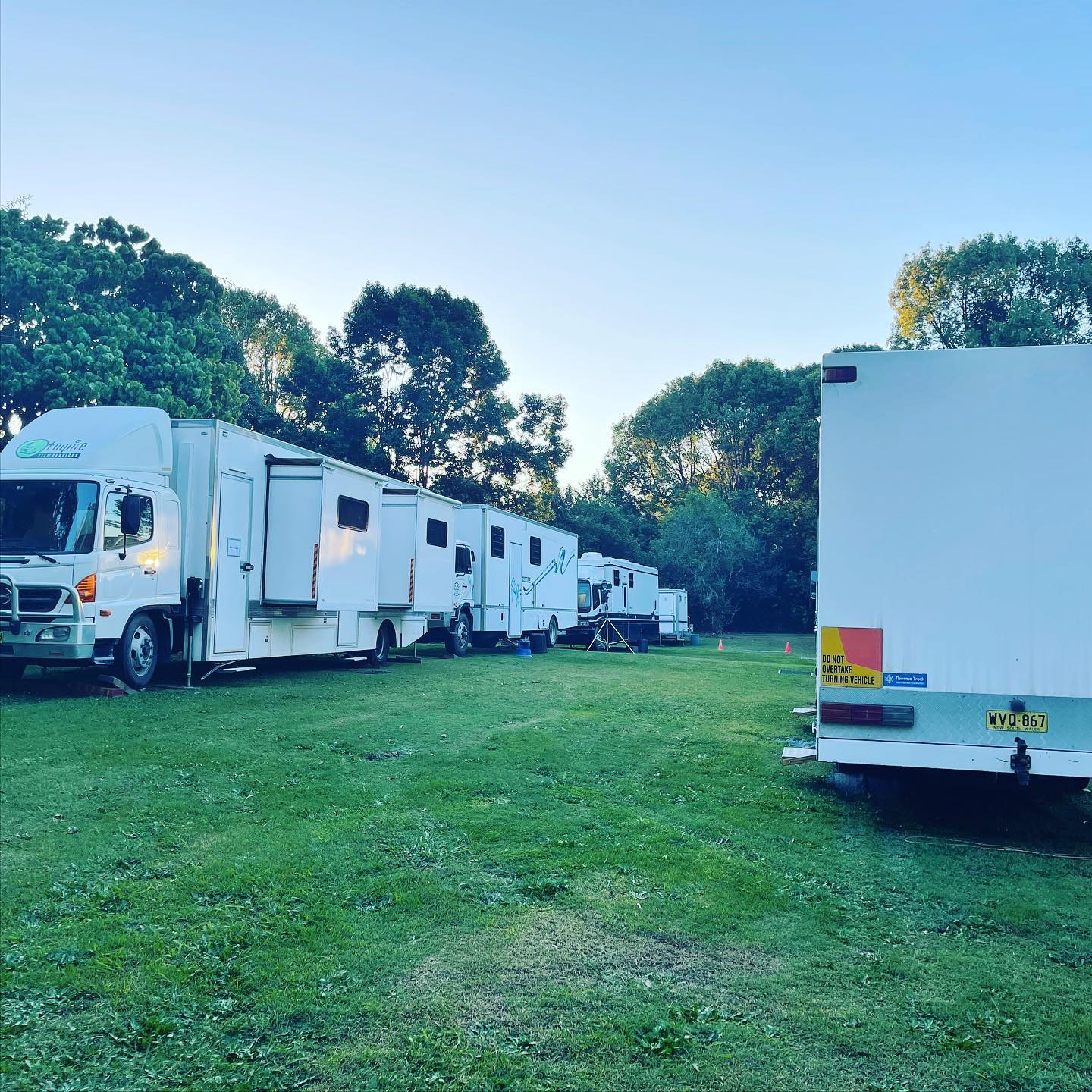 Row of white semi-trucks and trailers on green grass near trees under a blue sky, possibly a film or television production location.