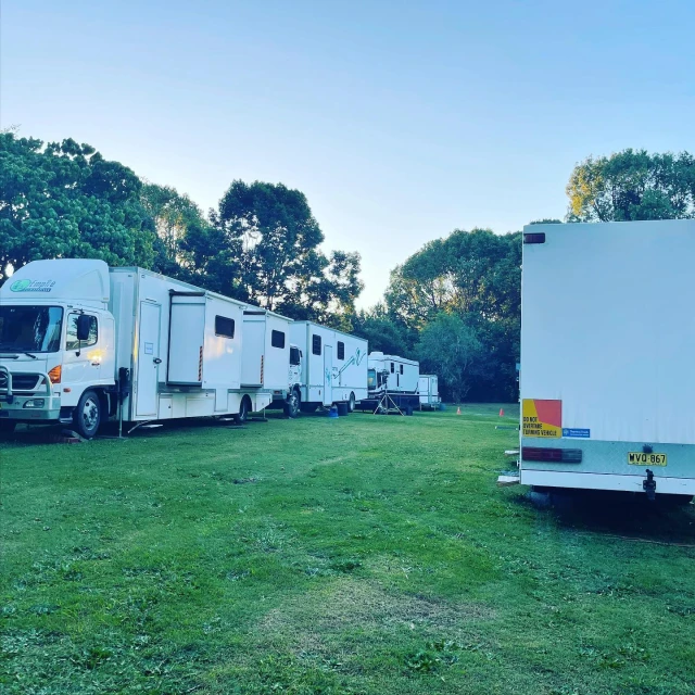 Row of white semi-trucks and trailers on green grass near trees under a blue sky, possibly a film or television production location.