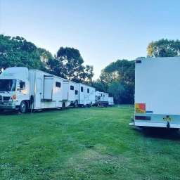 Row of white production trucks and trailers parked on a grassy field surrounded by trees under clear sky