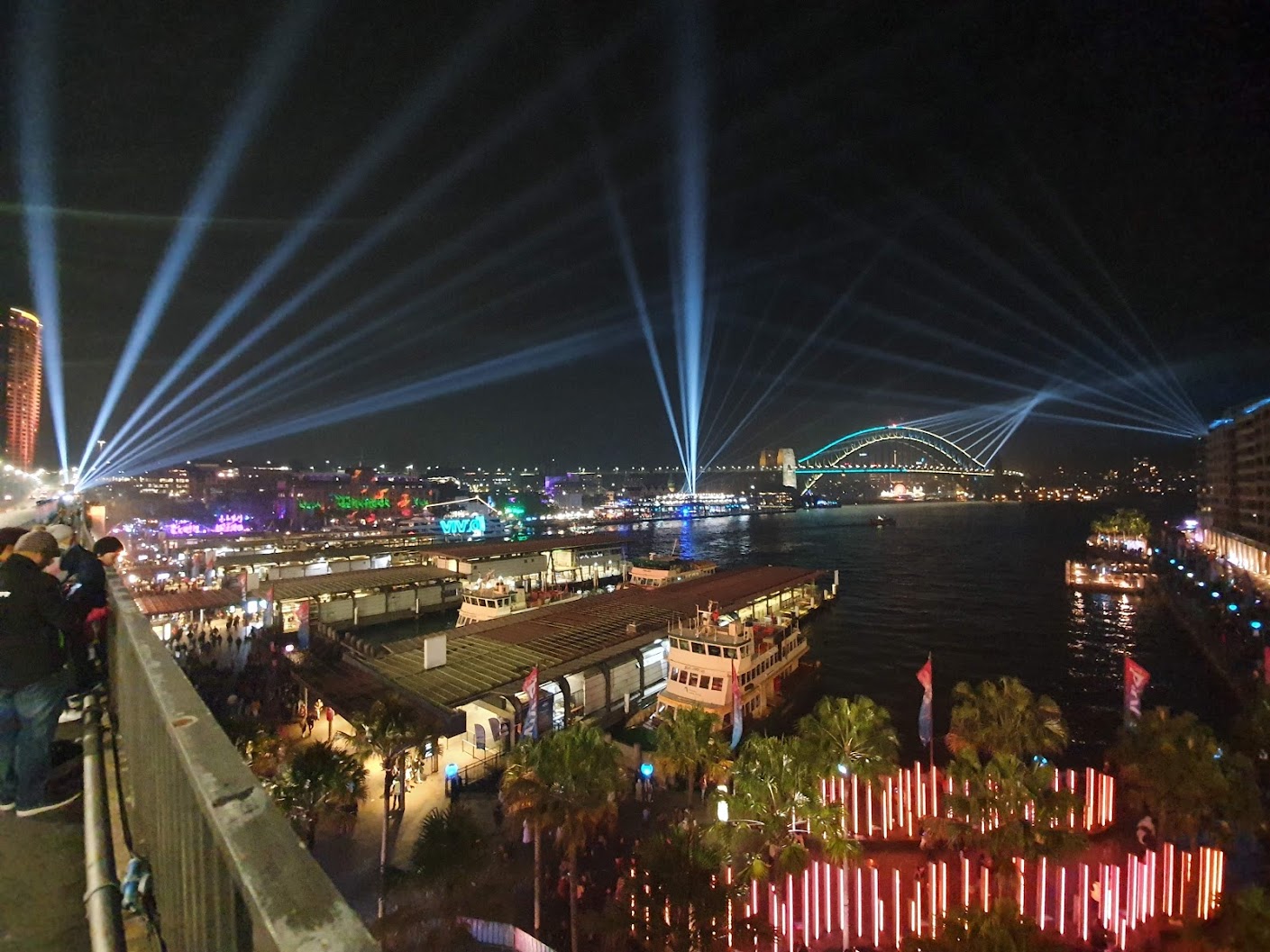 Night view of Darling Harbour in Sydney, Australia, featuring a bridge, buildings, bodies of water, light beams, and a crowd of people.